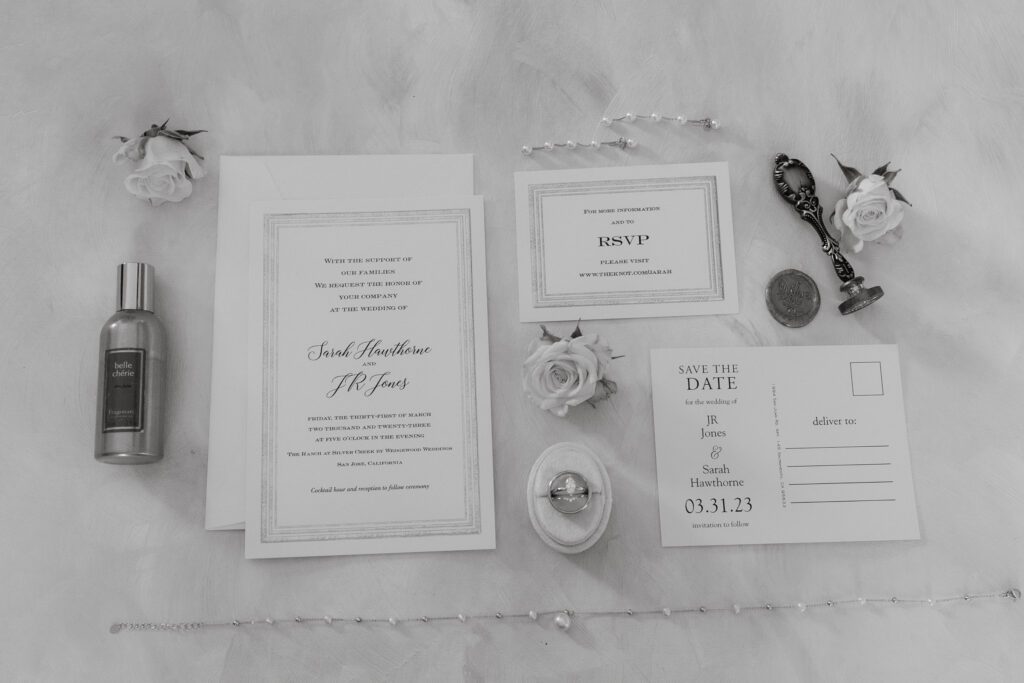 Black and white flatlay of wedding invitation suite with rings and florals