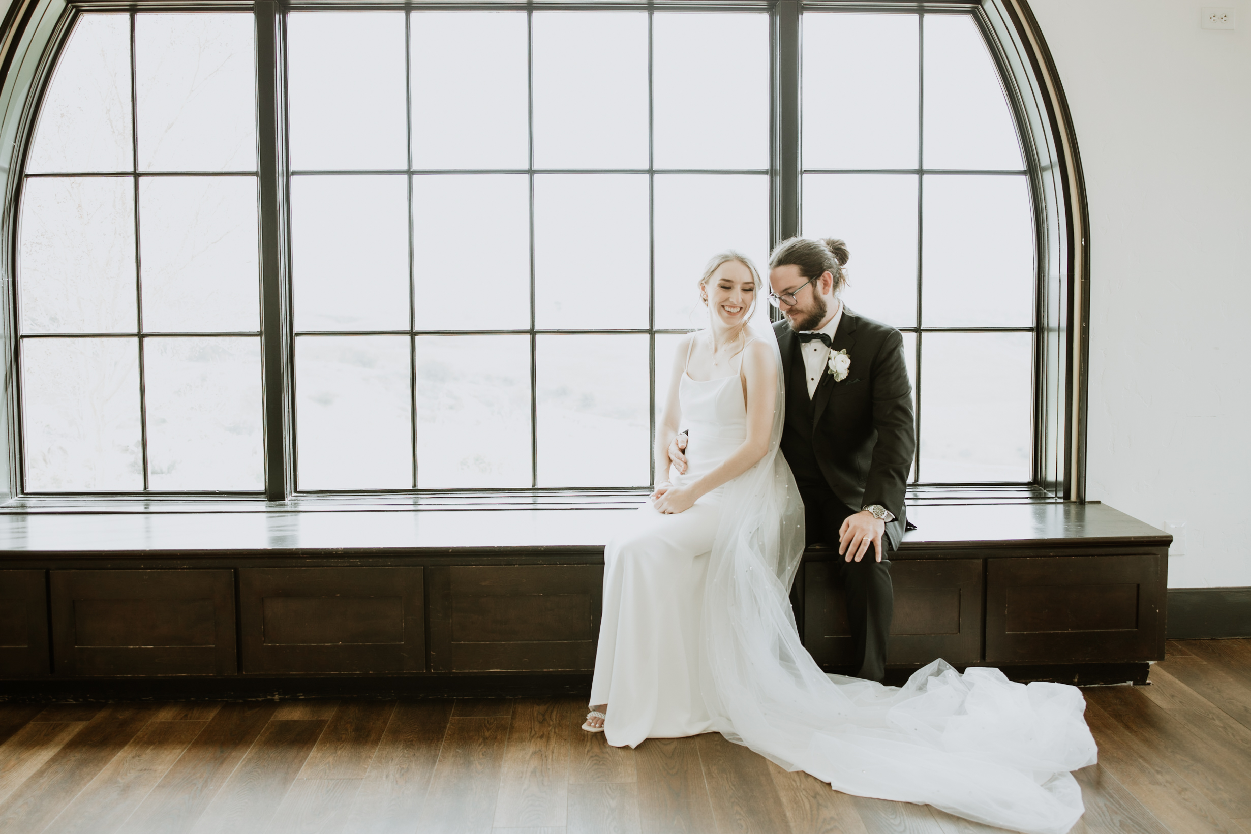 Bride and groom sitting by large arched window in elegant wedding portrait