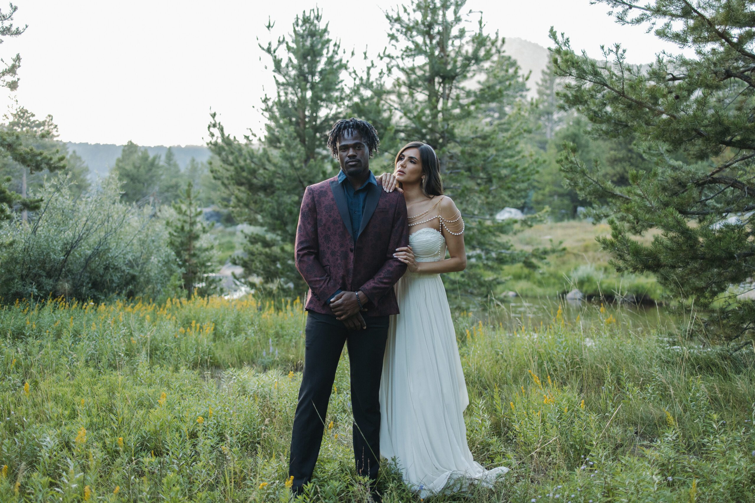 Man and Woman standing in a field of wildflowers with a mountain scape in the background. They are wearing bridal outfits looking at the camera.