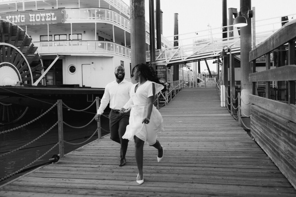 A black and white photo of a couple running towards the camera on a dock while looking at each other laughing