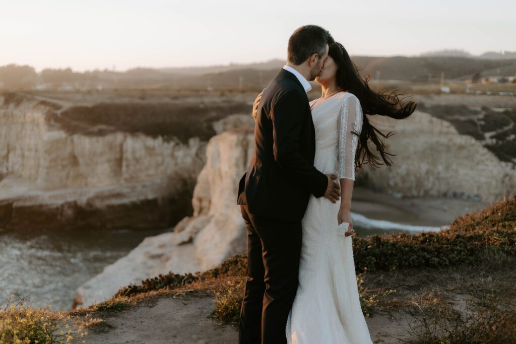 A newly wed couple standing by a coastal cliffside during a setting sun kissing romantically